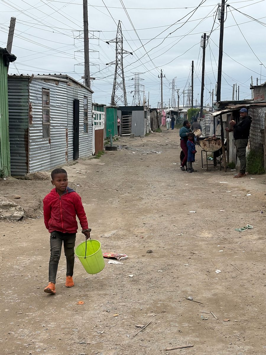 A young child in a red jacket carrying a green bucket along a street of corrugated-iron homes in Khayelitsha
